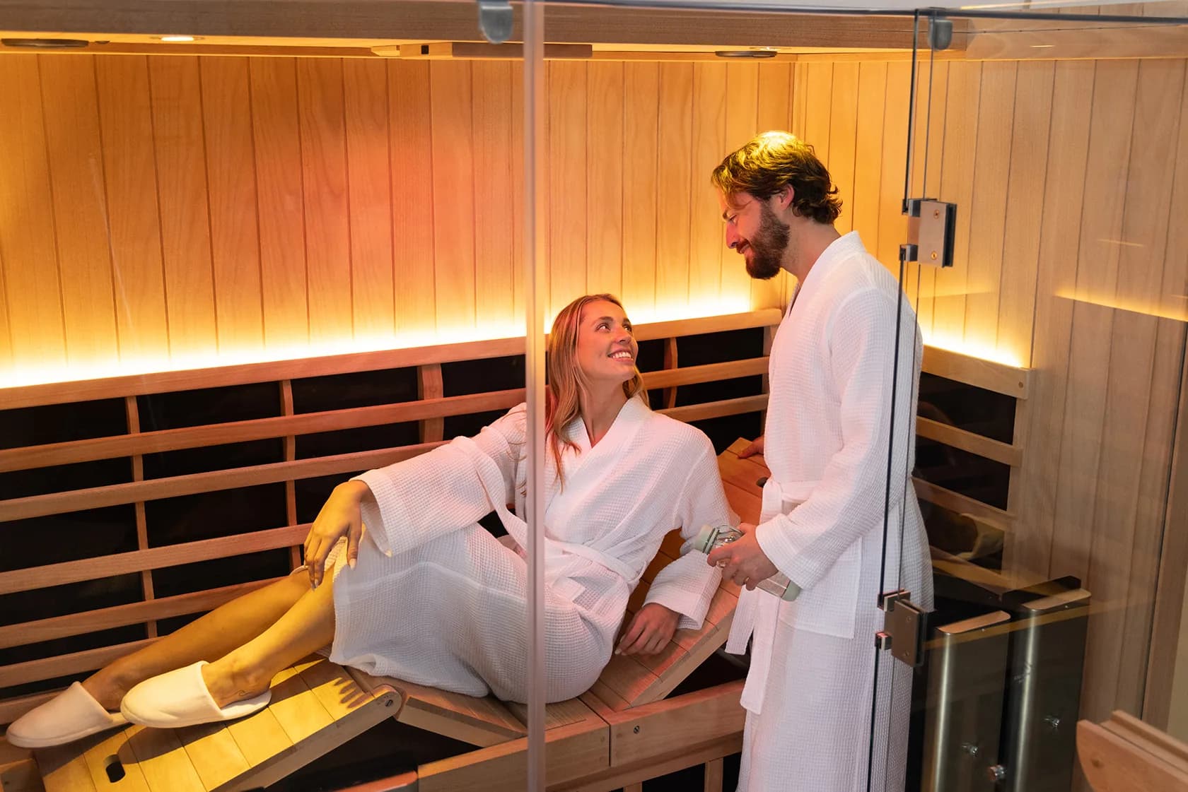 Clearlight's photograph of two people in white robes in a lit infrared cabin, one reclining on the bench and one standing at the glass door.