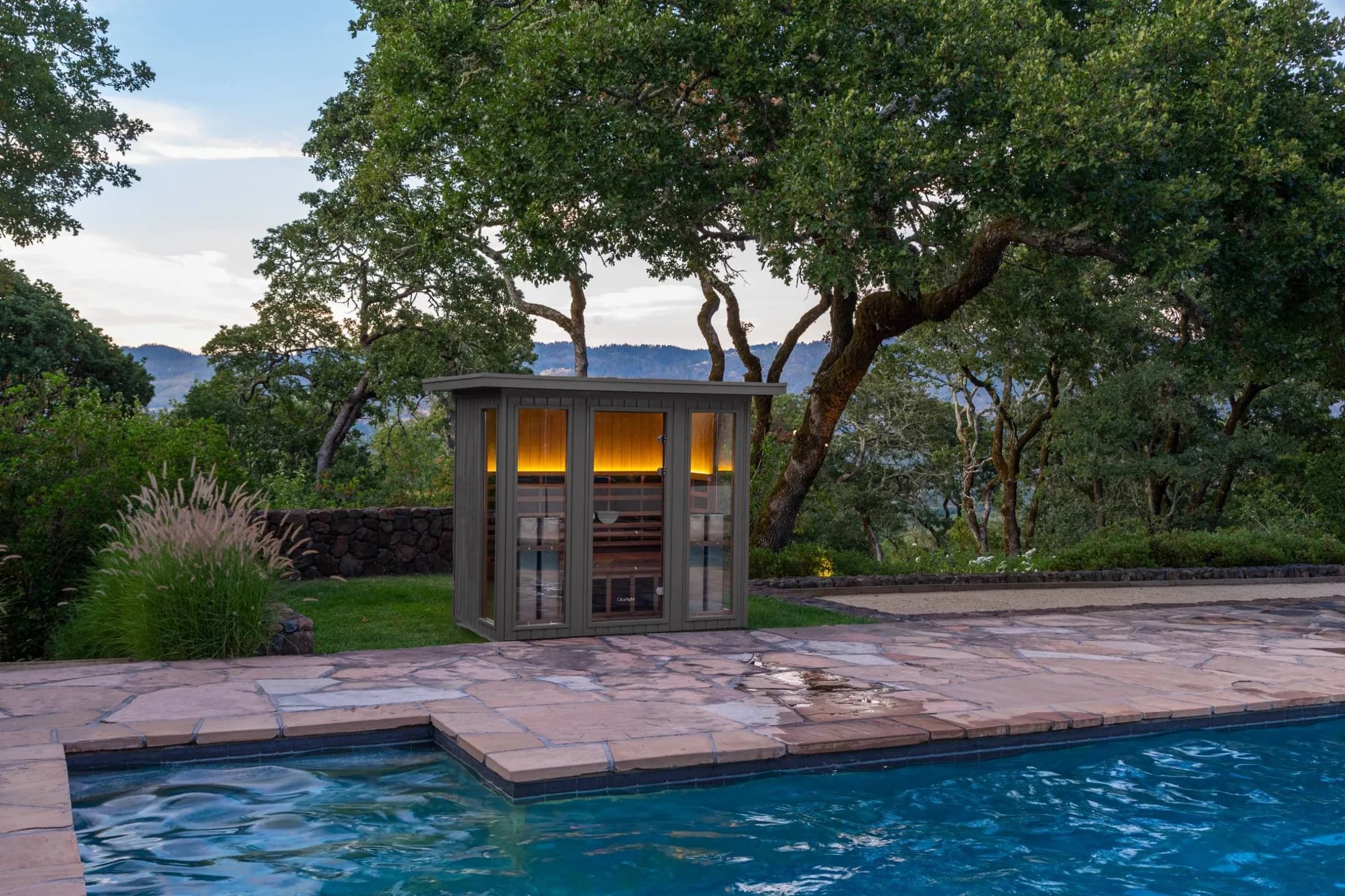Clearlight's own image of a Sanctuary Outdoor 5 cabin lit warm from inside at dusk, on a flagstone pool deck under oak trees with hills behind.