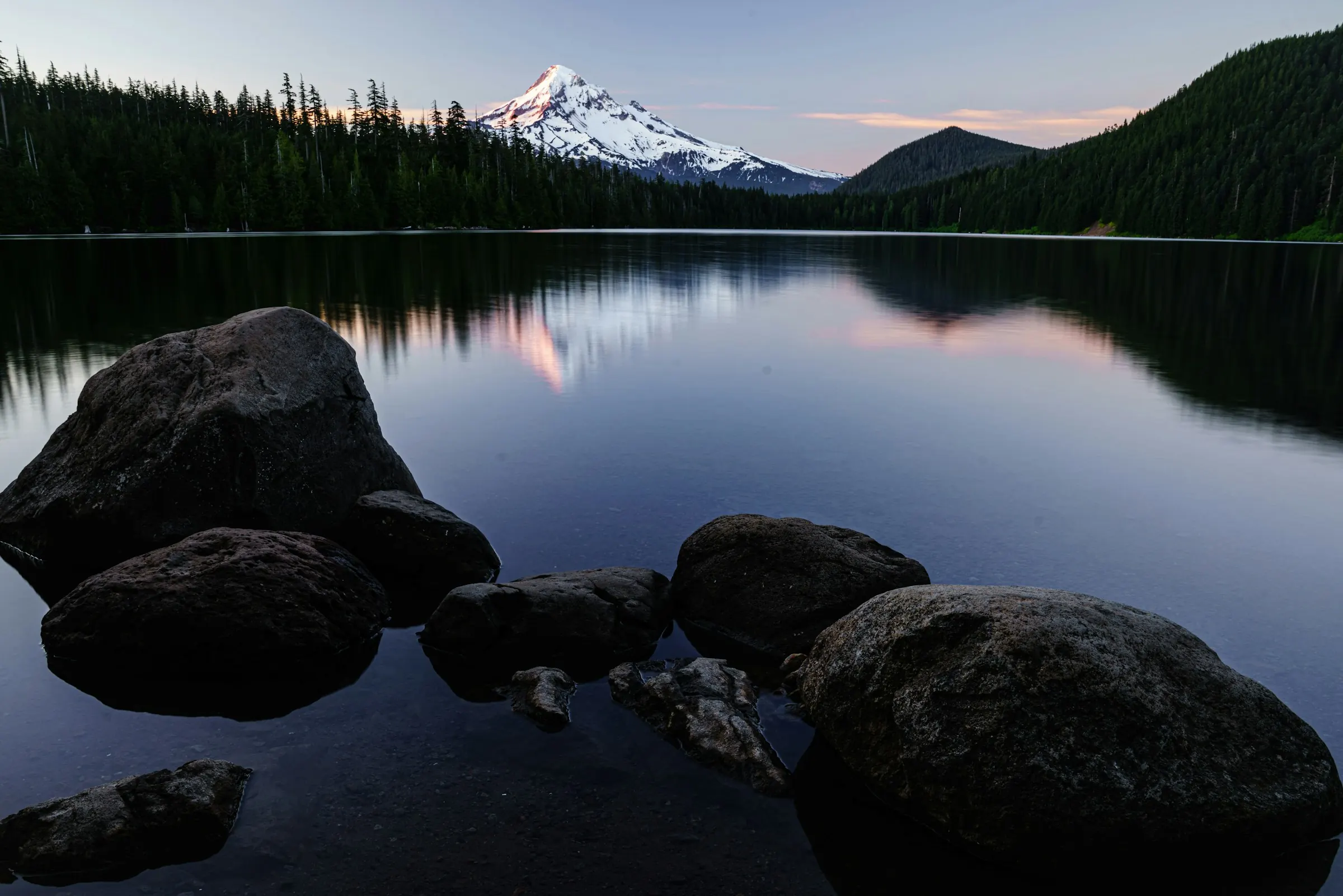 A still lake at dusk with boulders in the shallows, a pine shoreline, and a snow-capped peak beyond.