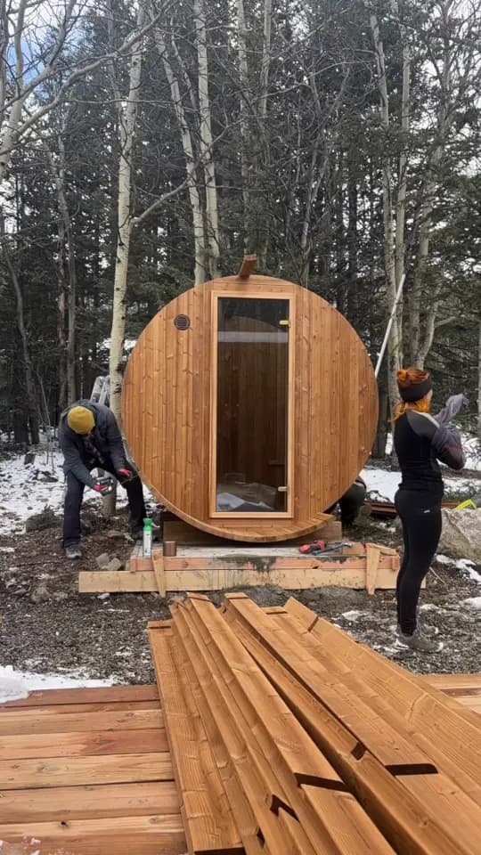 A frame from our time-lapse in Alma, Colorado: our crew assembling a barrel sauna on snow-patched ground among aspens and pines, its front wall and glass door standing and its staves stacked in the foreground.