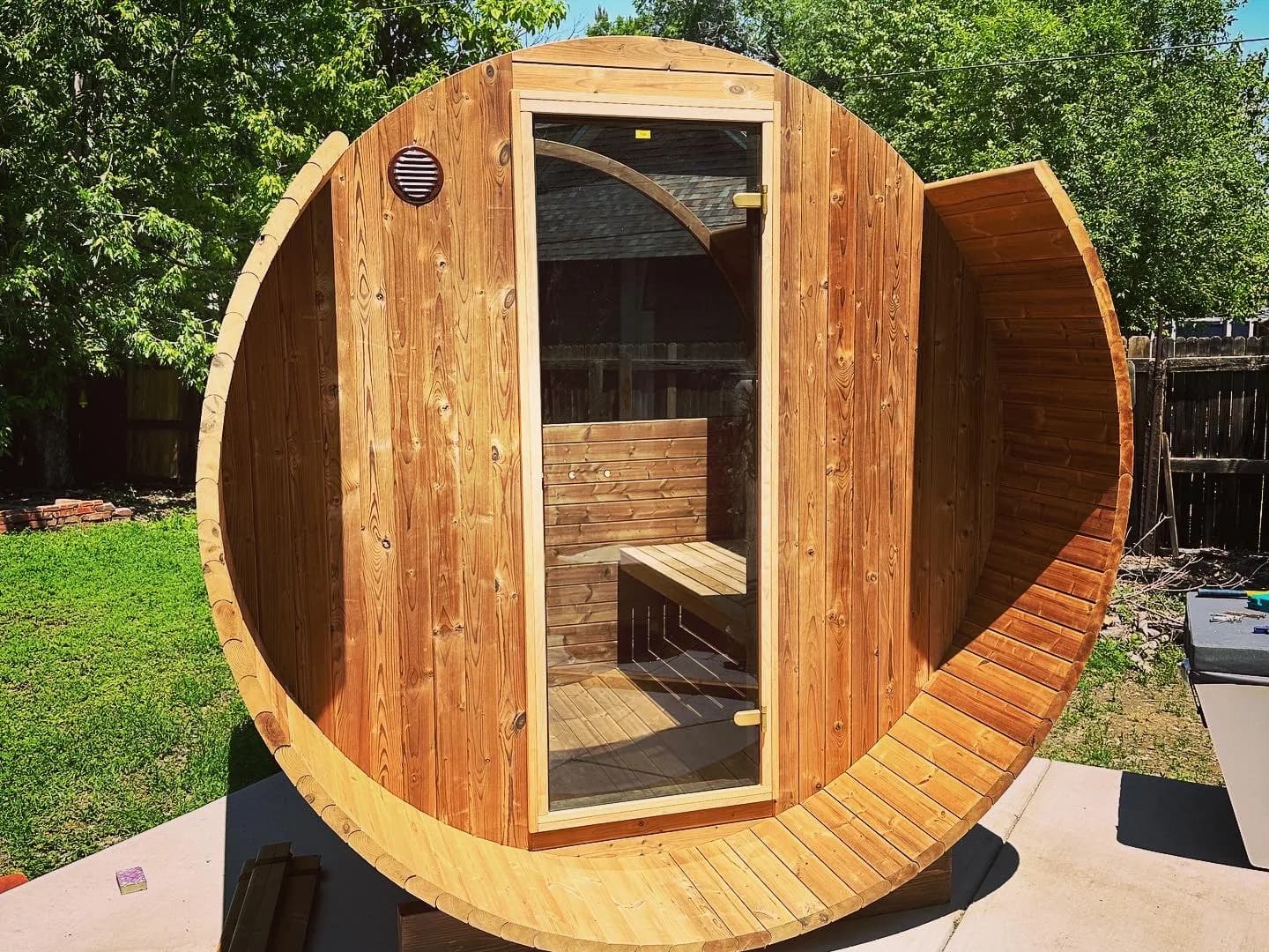 A cedar barrel sauna part way through assembly: the glass-door end wall standing, the staves up the sides behind it, and the top of the shell still open to the sky.