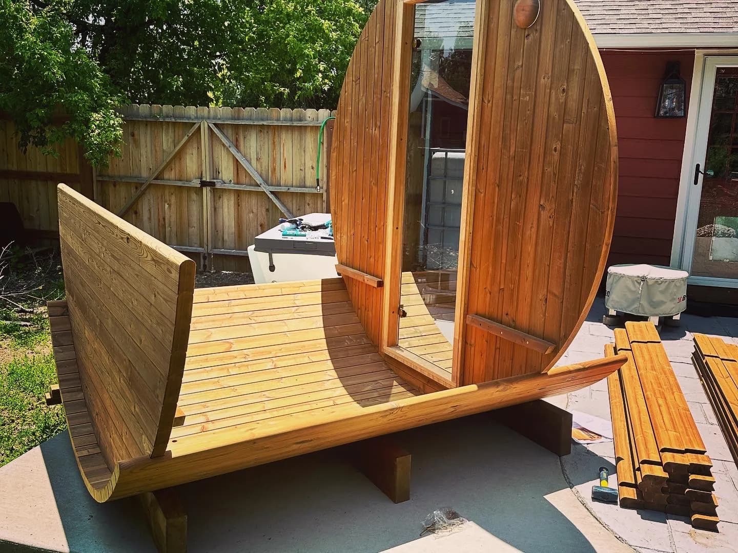 A barrel sauna part way through assembly on its cradles in a fenced back yard, the floor laid and the front wall standing.