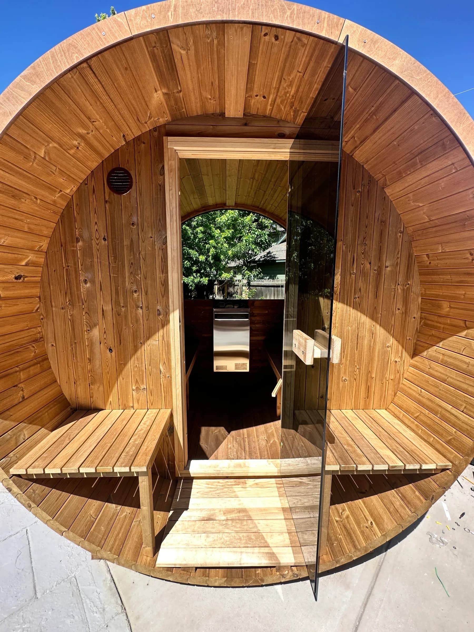 A cedar barrel sauna with its glass door open, benches on the front porch, and the interior visible through to a rear window onto trees. One of our Colorado builds.