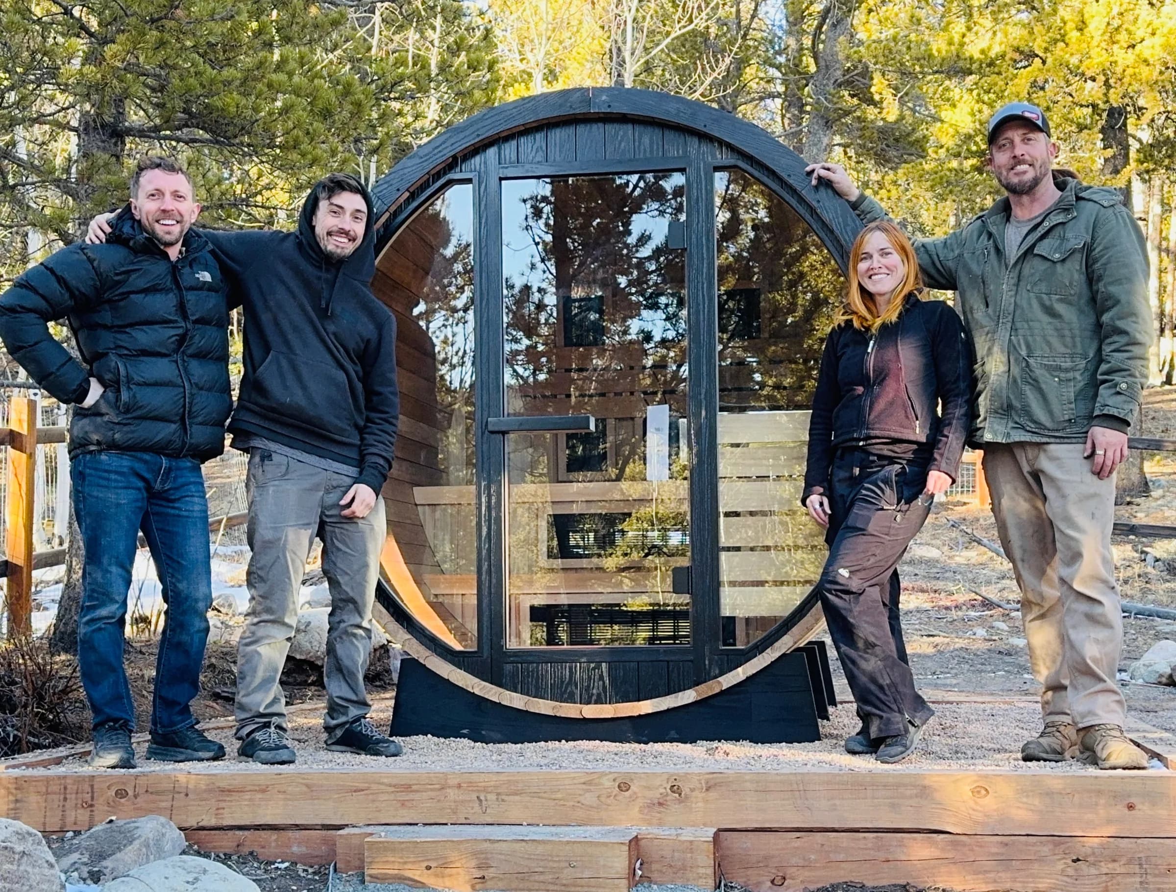 Two members of our crew standing either side of a finished black barrel sauna with a round glass front, on a timber-framed gravel base among pines and aspens.
