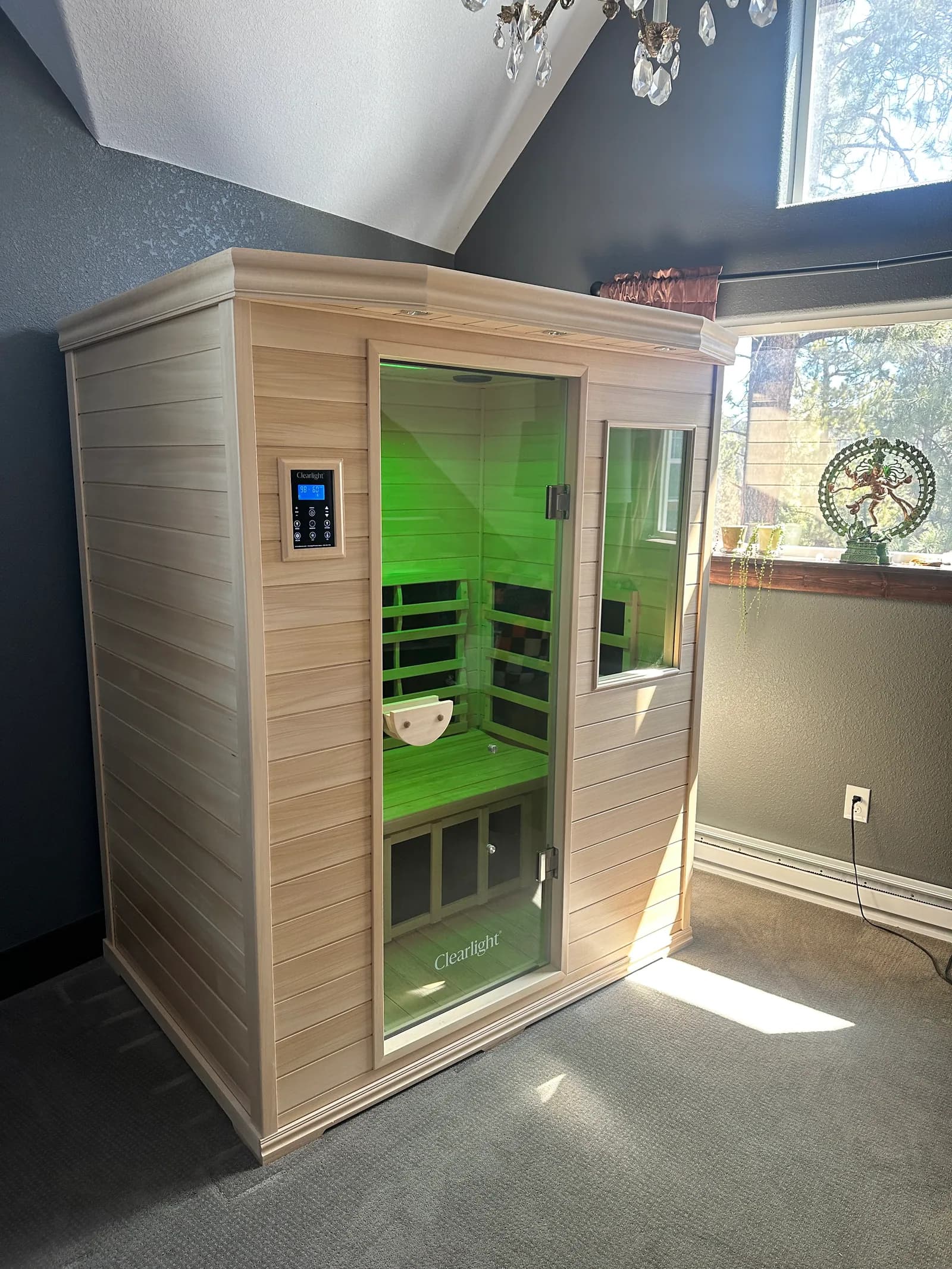 A Clearlight infrared cabin in a carpeted room beside a sunny window, its interior lit green behind the glass door.