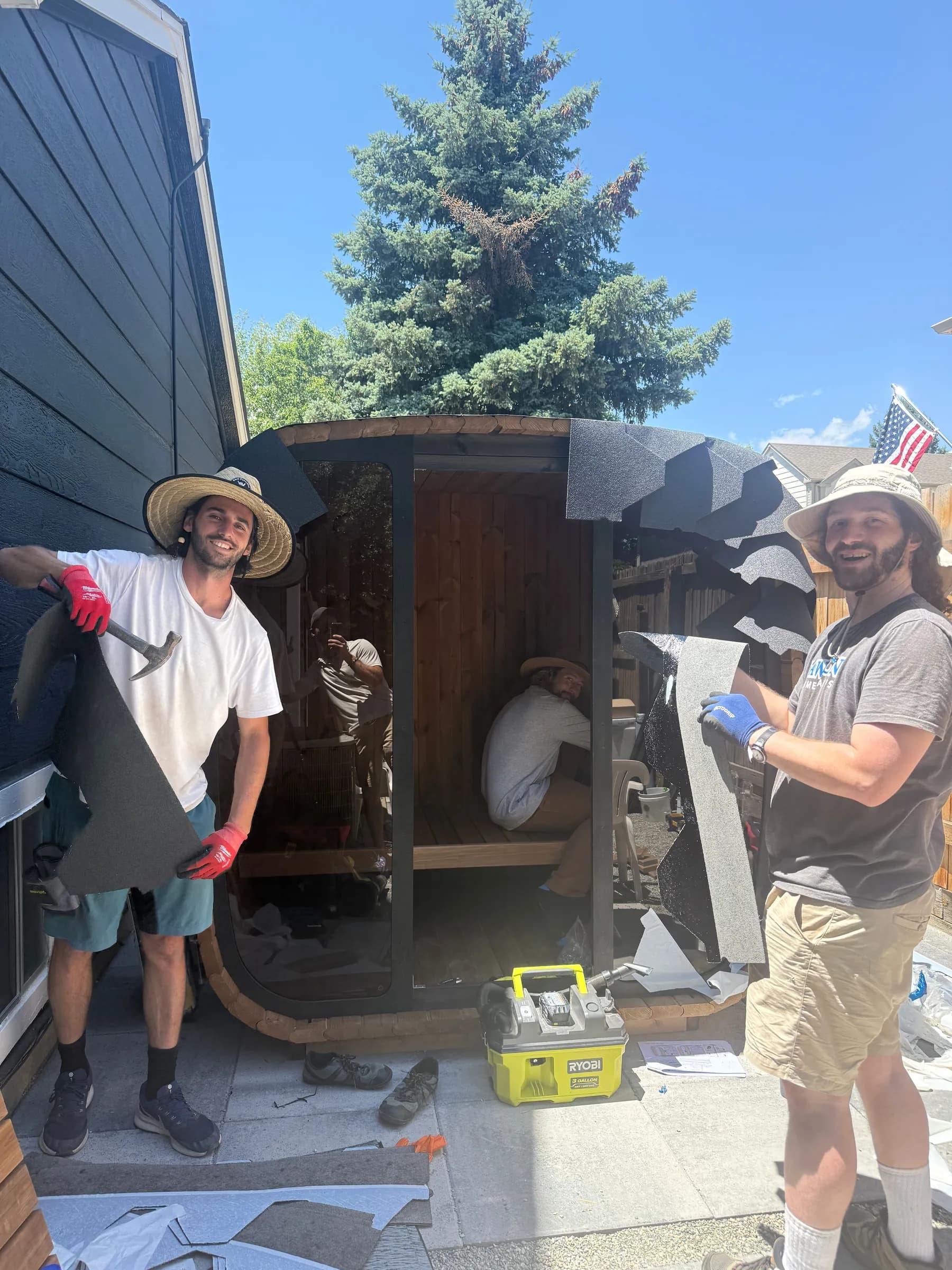 Two of our crew in gloves fitting the roofing on a dark cube sauna beside a house, a pine tree behind.